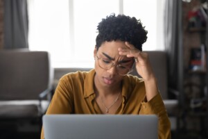 Black woman appearing tired and frustrated holding head with hand while sitting in front of laptop