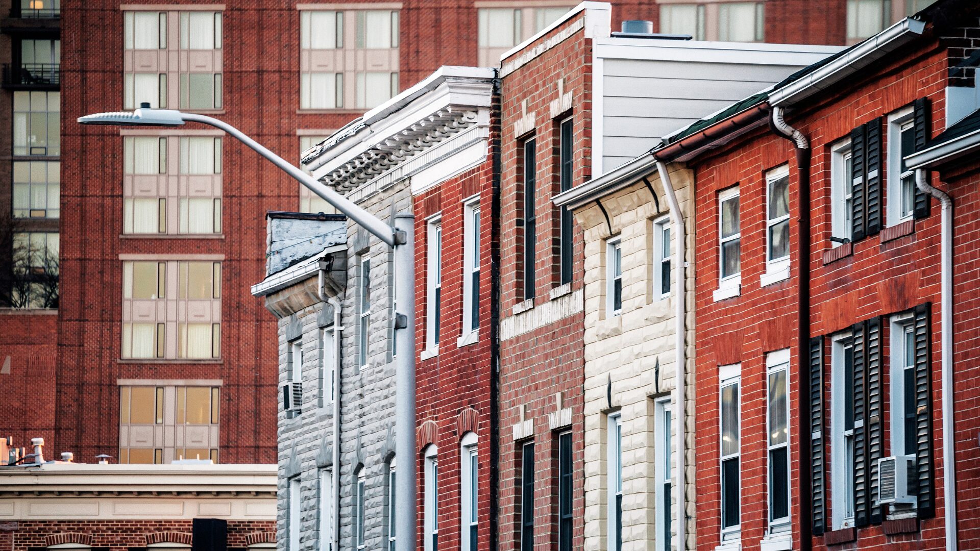 Row of attached brick and stone rowhouses along a city street, with varied facades, windows, and a streetlight in the foreground against taller buildings in the background.