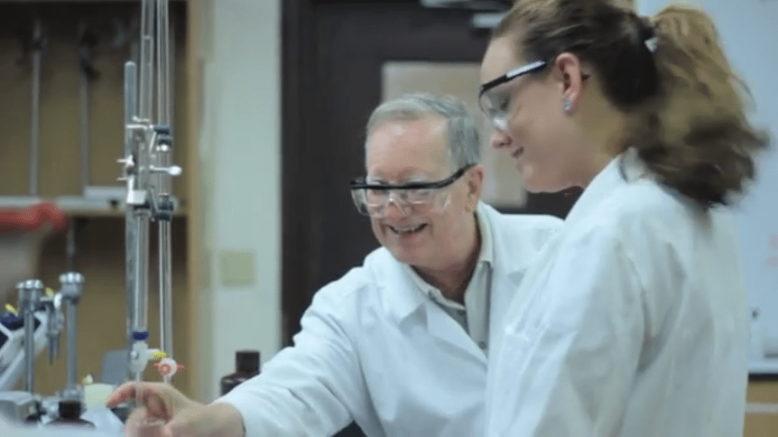 A male chemistry teacher and a female student wearing white lab coats and safety goggles smile while working together in a science laboratory.