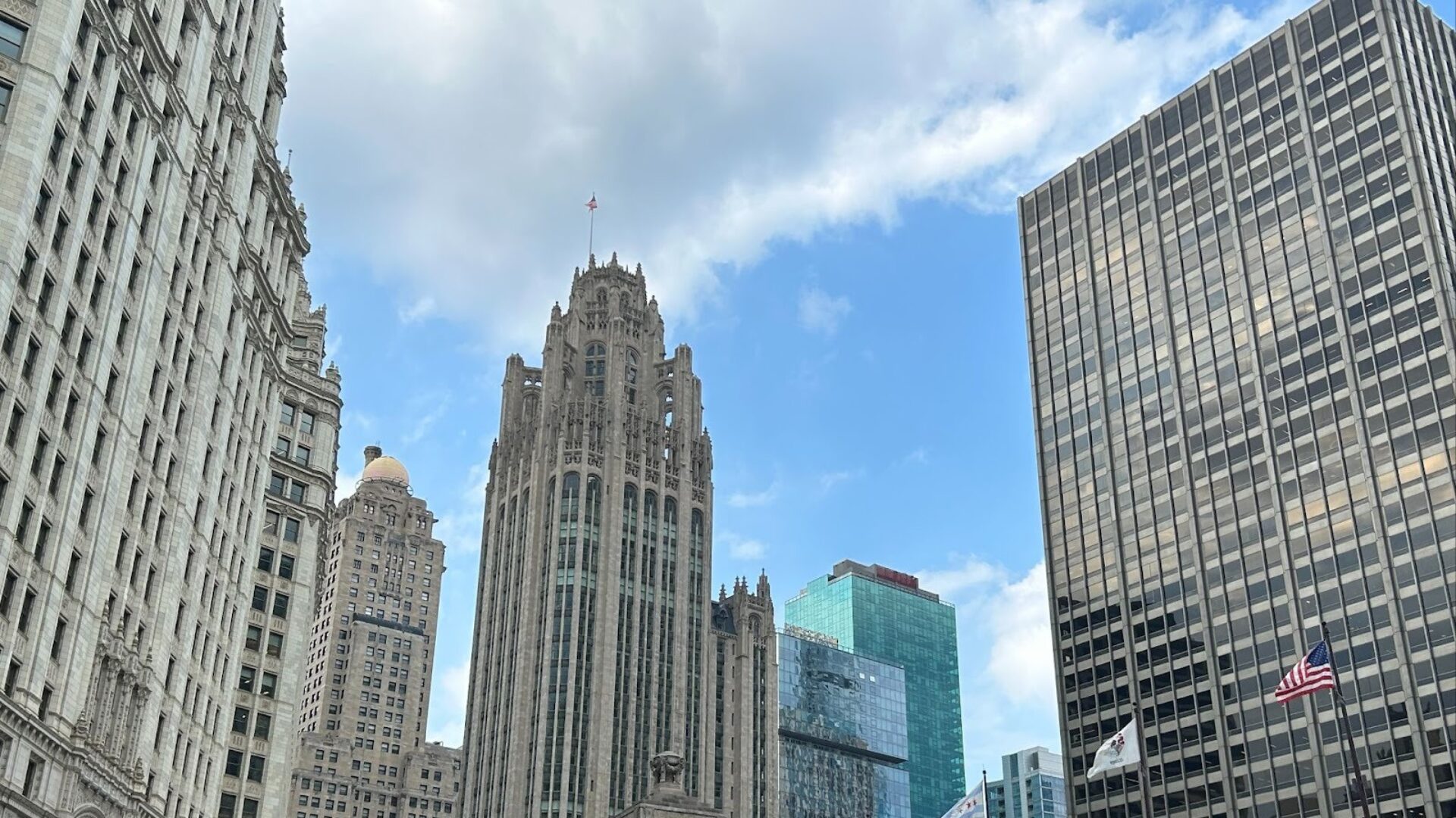 Downtown city skyline with historic and modern high-rise buildings, including a tall gothic-style tower, glass office buildings, and American flags under a partly cloudy sky.
