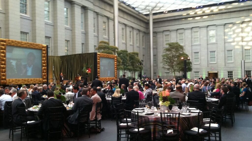 A conference filmed by minimatters with many people sitting at round tables arranged in rows with a stage in the back
