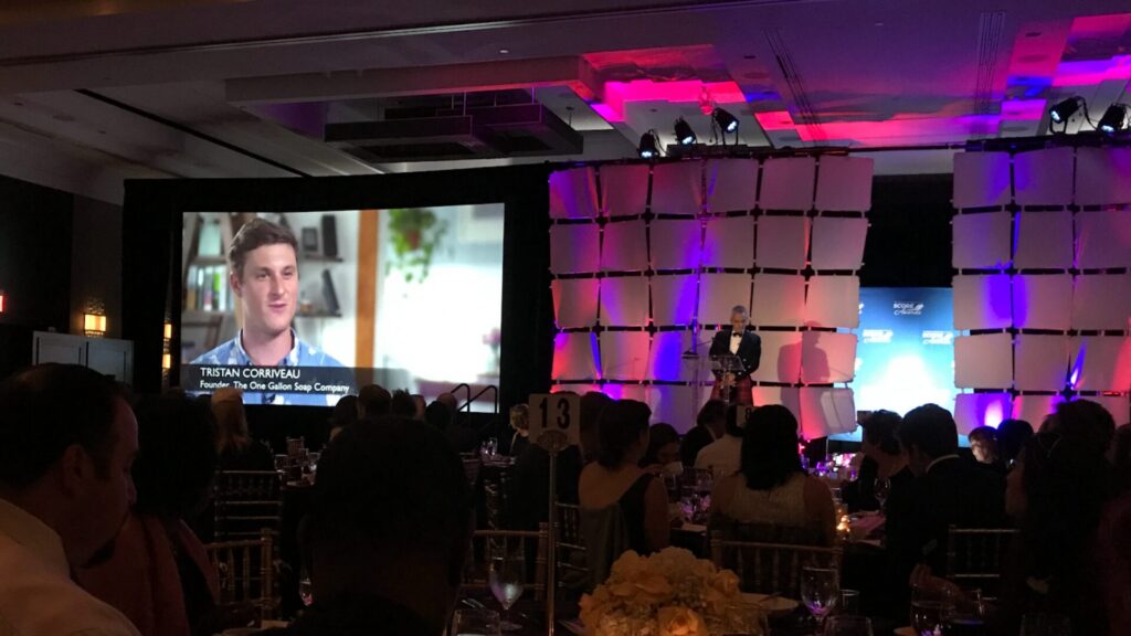 A gala filmed by MiniMatters with a bunch of people sitting in a big room. Low colorful lights and a presentation being shown on the screen in the front