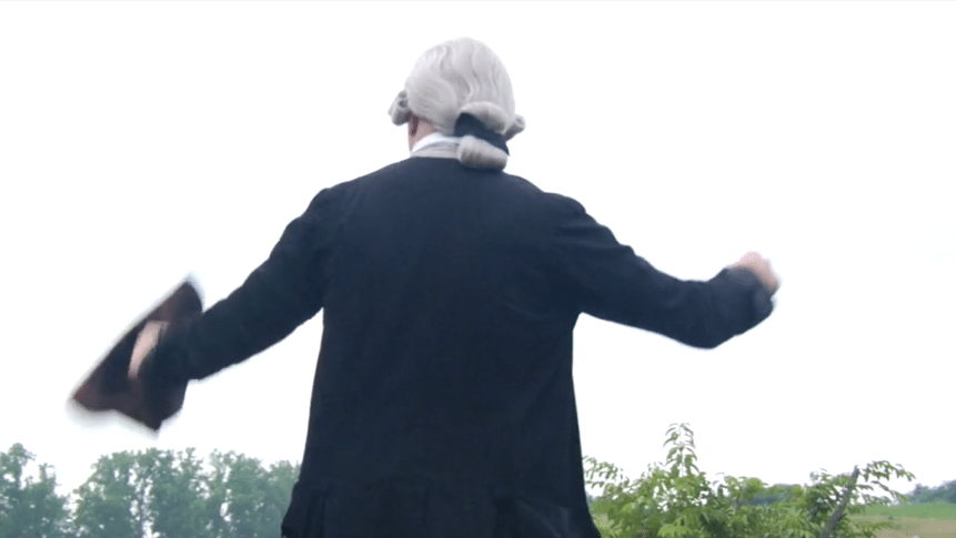 Rear view of a historical reenactor dressed as George Washington, wearing a powdered wig and dark coat, overlooking a green landscape.