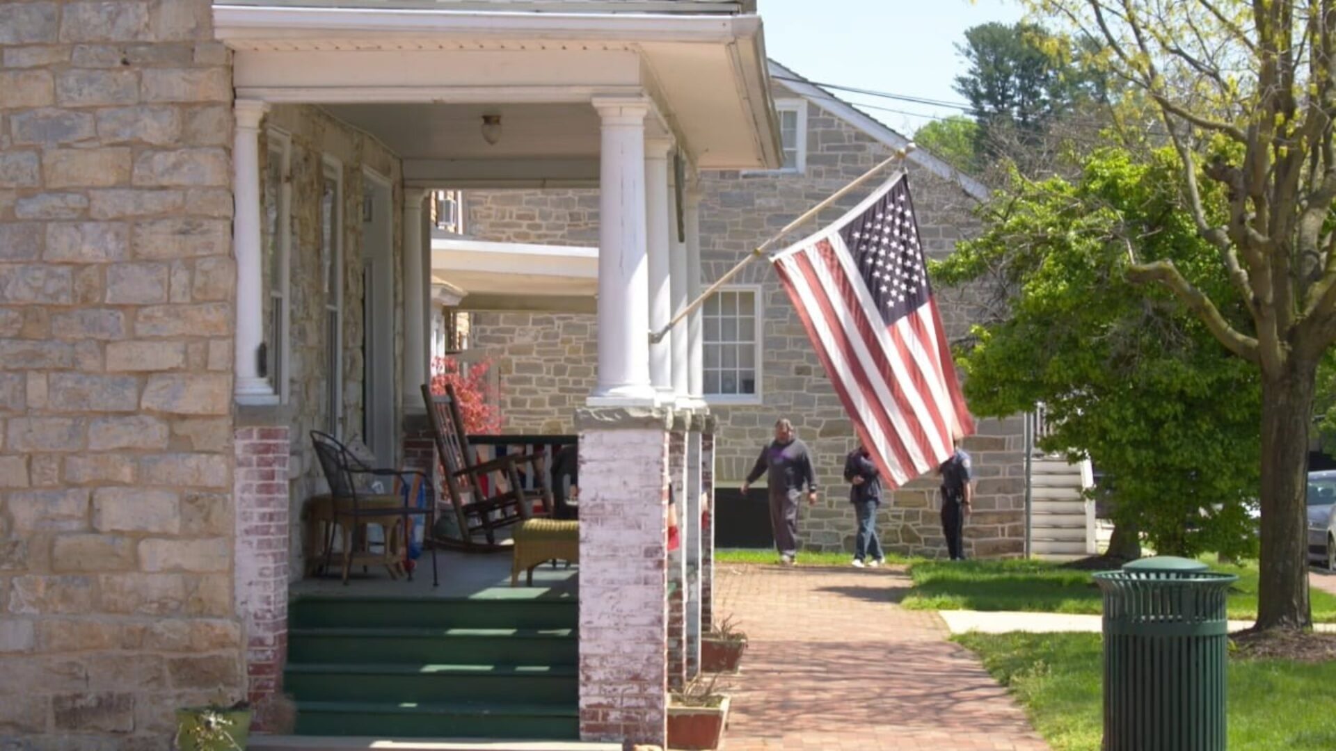 Front porch of a stone house with rocking chairs and a large American flag hanging beside a brick walkway, with people walking in the background on a sunny day.