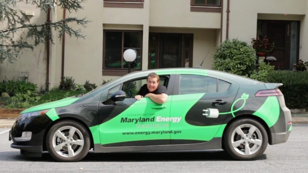 Person leaning out of the driver’s window of a green Maryland Energy–branded electric vehicle parked outside a residential building.