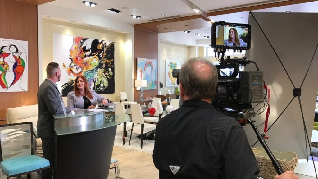 Behind-the-scenes view of a video production shoot, with a camera operator filming an interview between two people in a modern office setting.