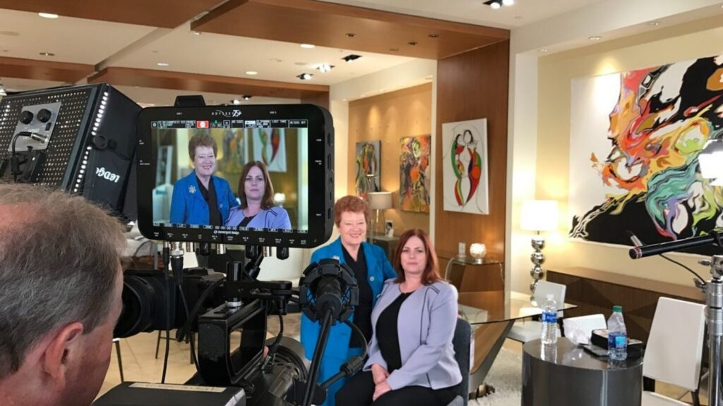 Camera filming two women seated in a modern office setting, with their interview framed on a monitor while lighting and production equipment are set up around them.
