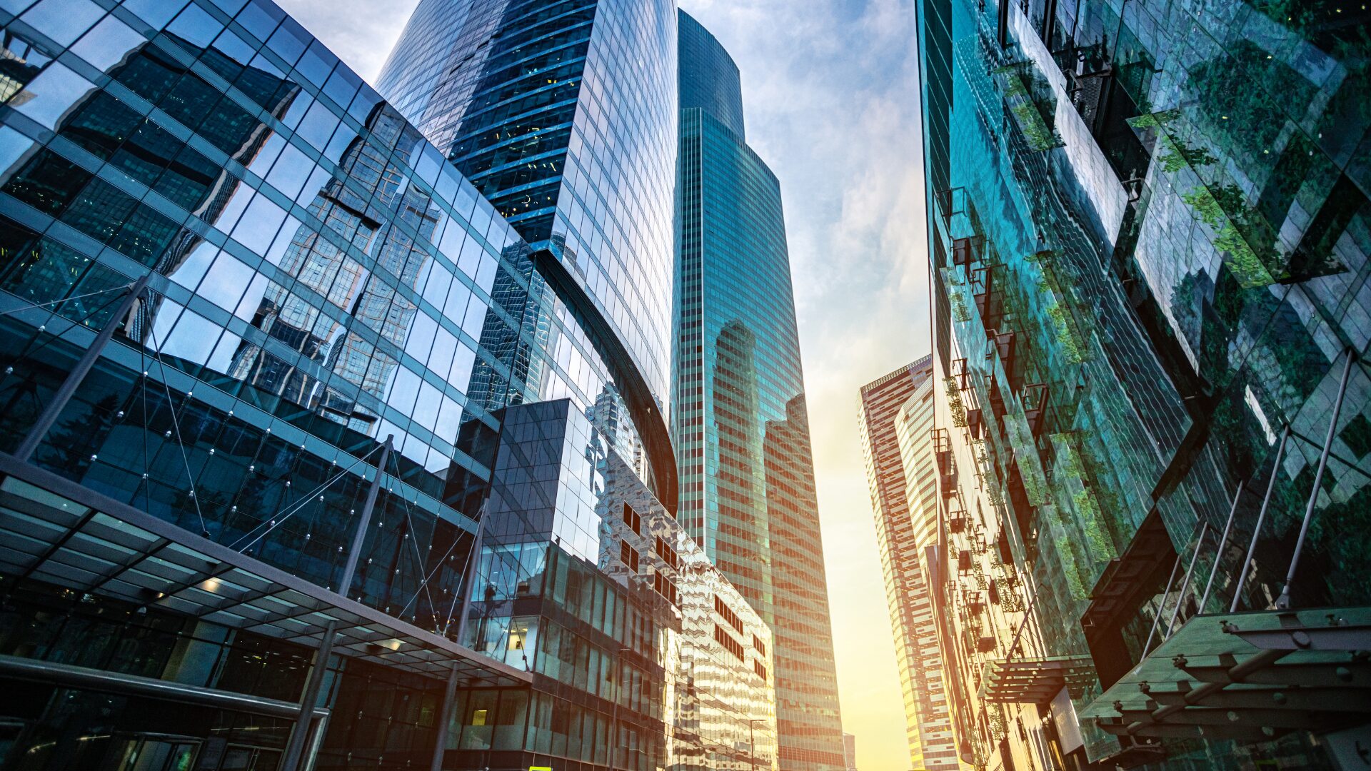 Modern glass skyscrapers reflecting the sky and surrounding city, viewed from street level in a downtown business district.