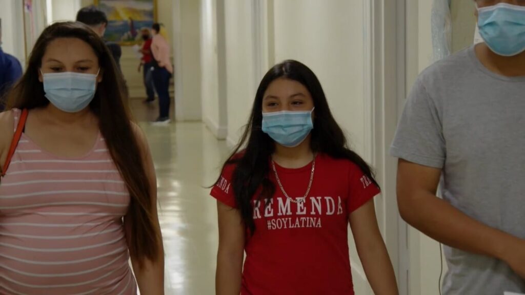 A mother on the left with her daughter in the middle, both wearing masks and walking in a hallway