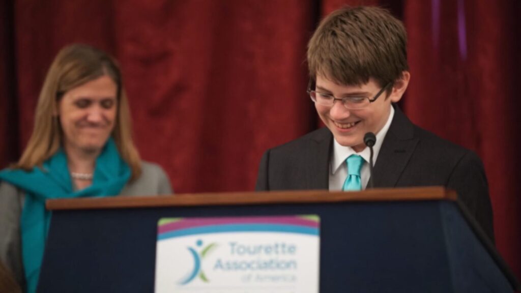 A man standing behind a podium looking down, with a women in the background