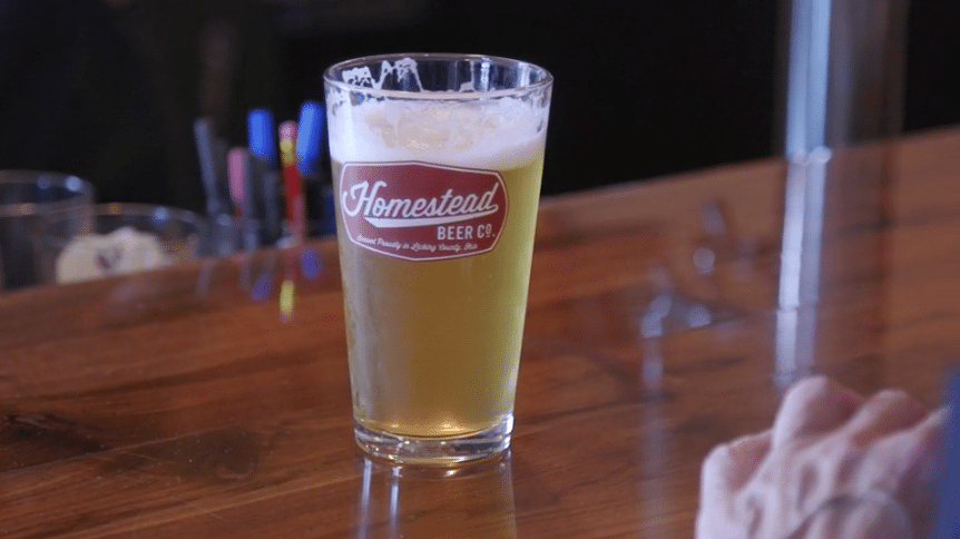 A pint glass of light-colored beer with the Homestead Beer Co. logo on a wooden bar top.