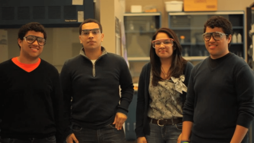 Four high school chemistry students wearing safety goggles standing together in a science laboratory.