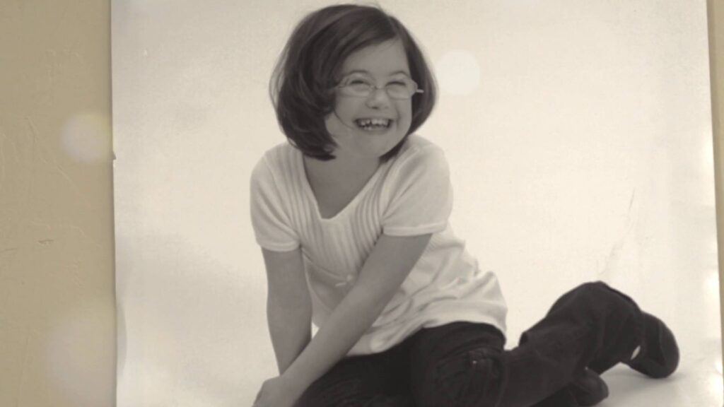 A young girl with glasses and short hair laughing while sitting on a white floor in a black-and-white studio portrait.