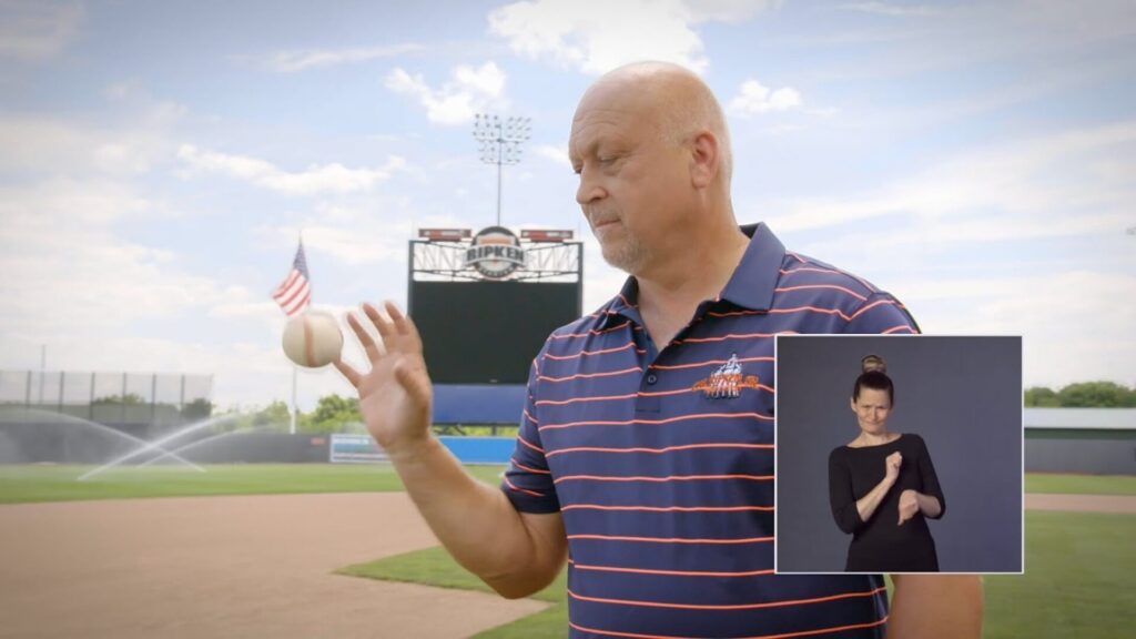 Man standing on a baseball field spinning a baseball in his hand, with a stadium scoreboard and American flag in the background, and a sign language interpreter shown in an inset on the screen.
