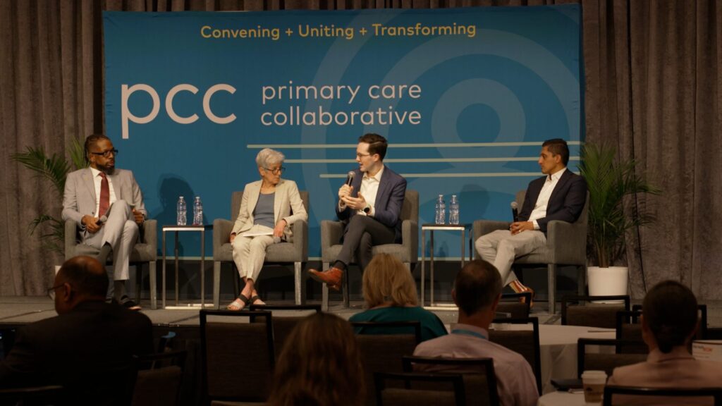 Panel discussion at a Primary Care Collaborative conference, with four speakers seated on stage addressing an audience.