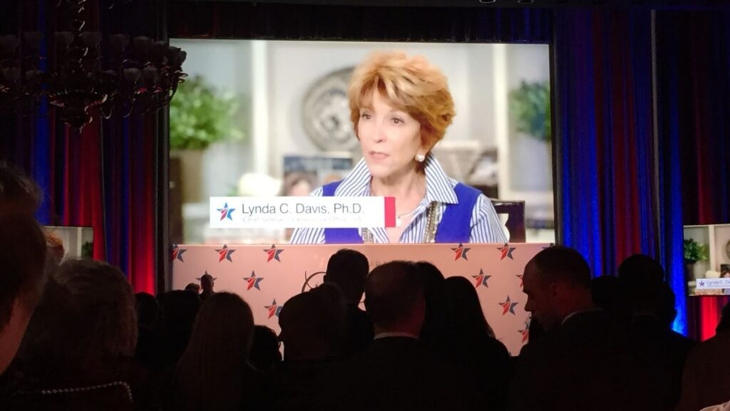 Audience watching a keynote presentation on a large screen featuring a speaker identified as Lynda C. Davis, Ph.D., at a live conference event.