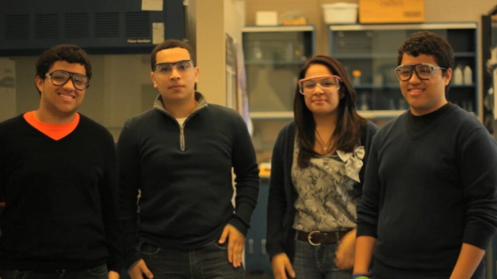Four students wearing safety goggles standing together in a laboratory setting.