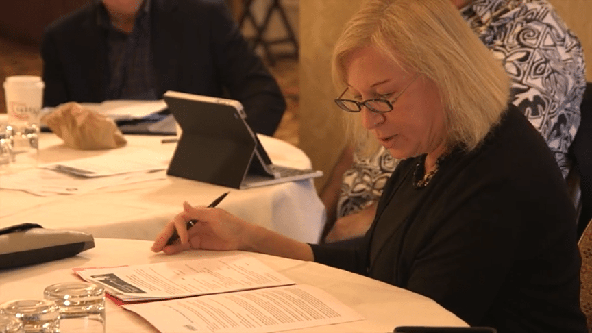 A blonde woman with glasses intently reading a document while participating in a professional think tank session.