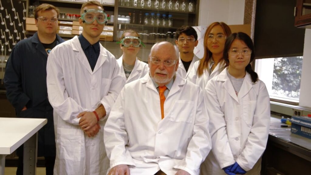 Multiple people standing in a room looking at the camera. They are all wearing lab coats.