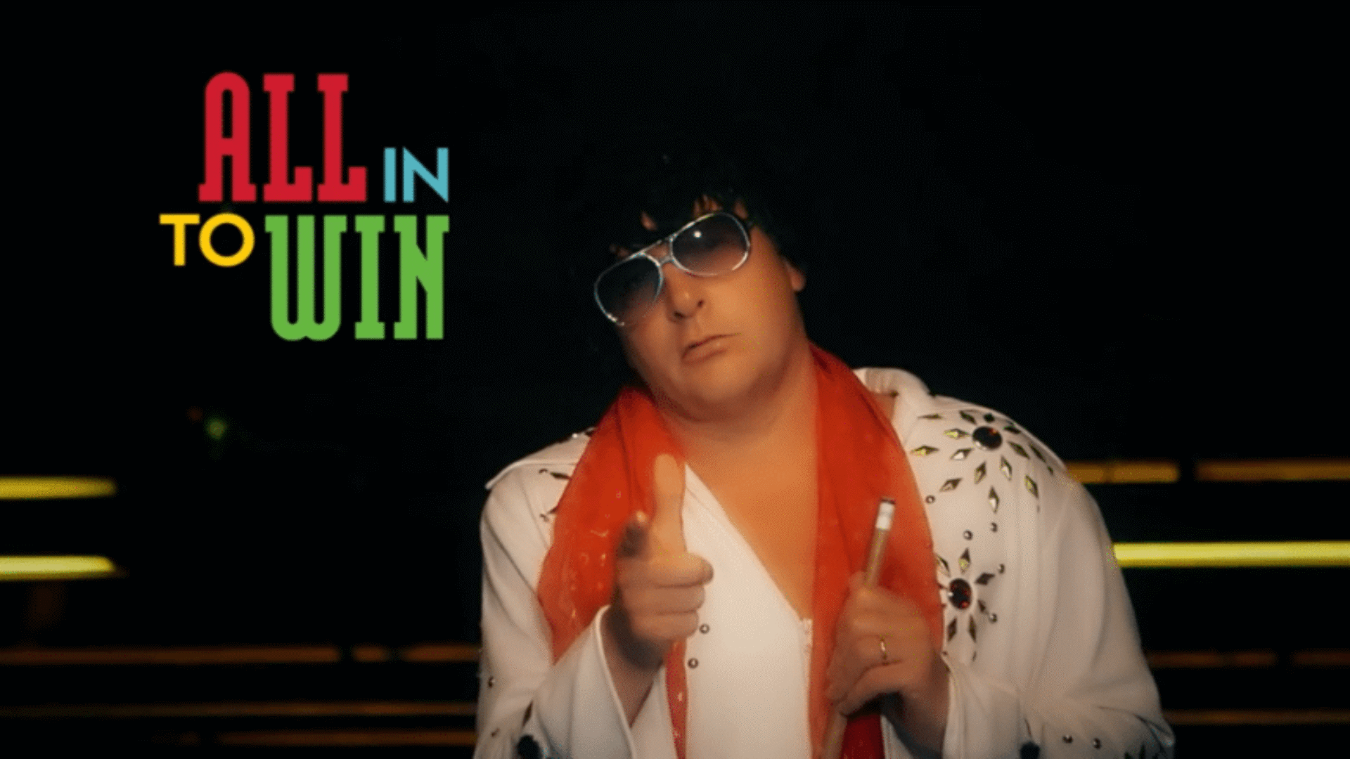 Person dressed in an Elvis-style costume pointing toward the camera with text “All In to Win” on the left.