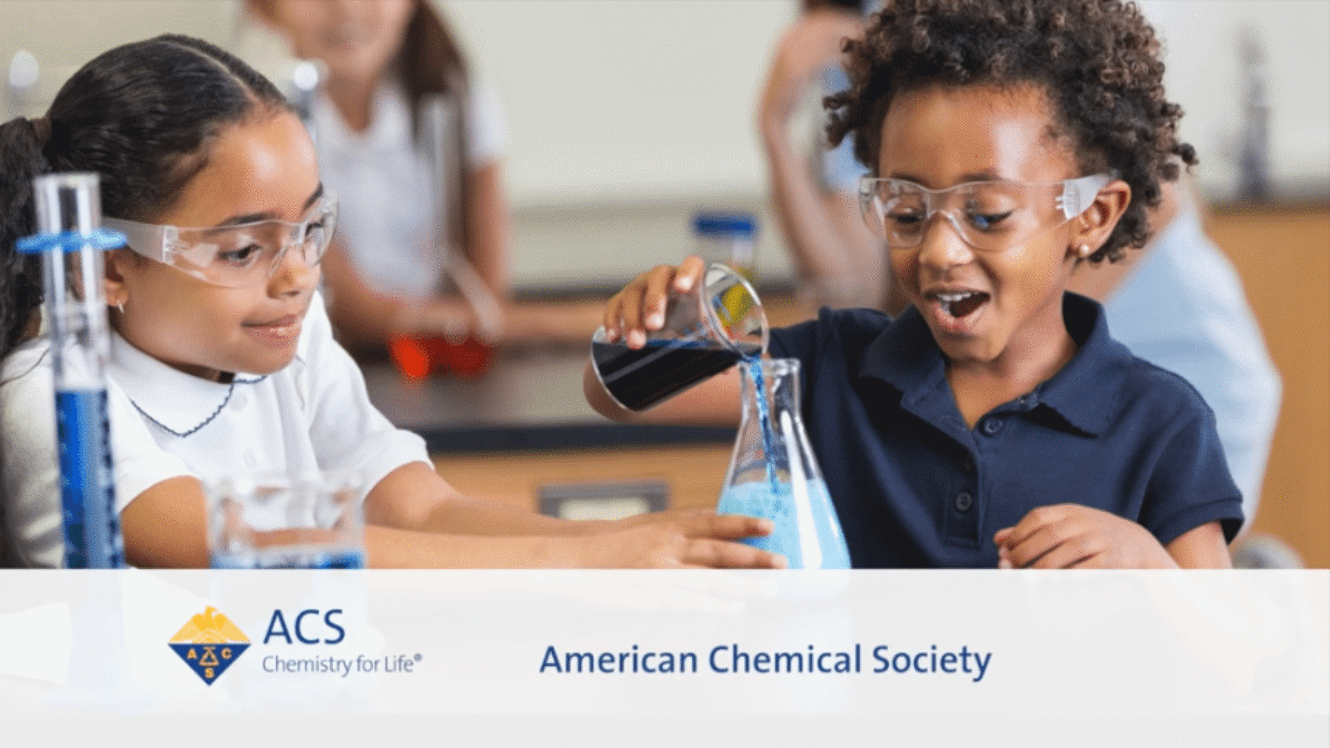 Two young students wearing safety goggles smile while conducting a chemistry experiment for an American Chemical Society (ACS) educational video.
