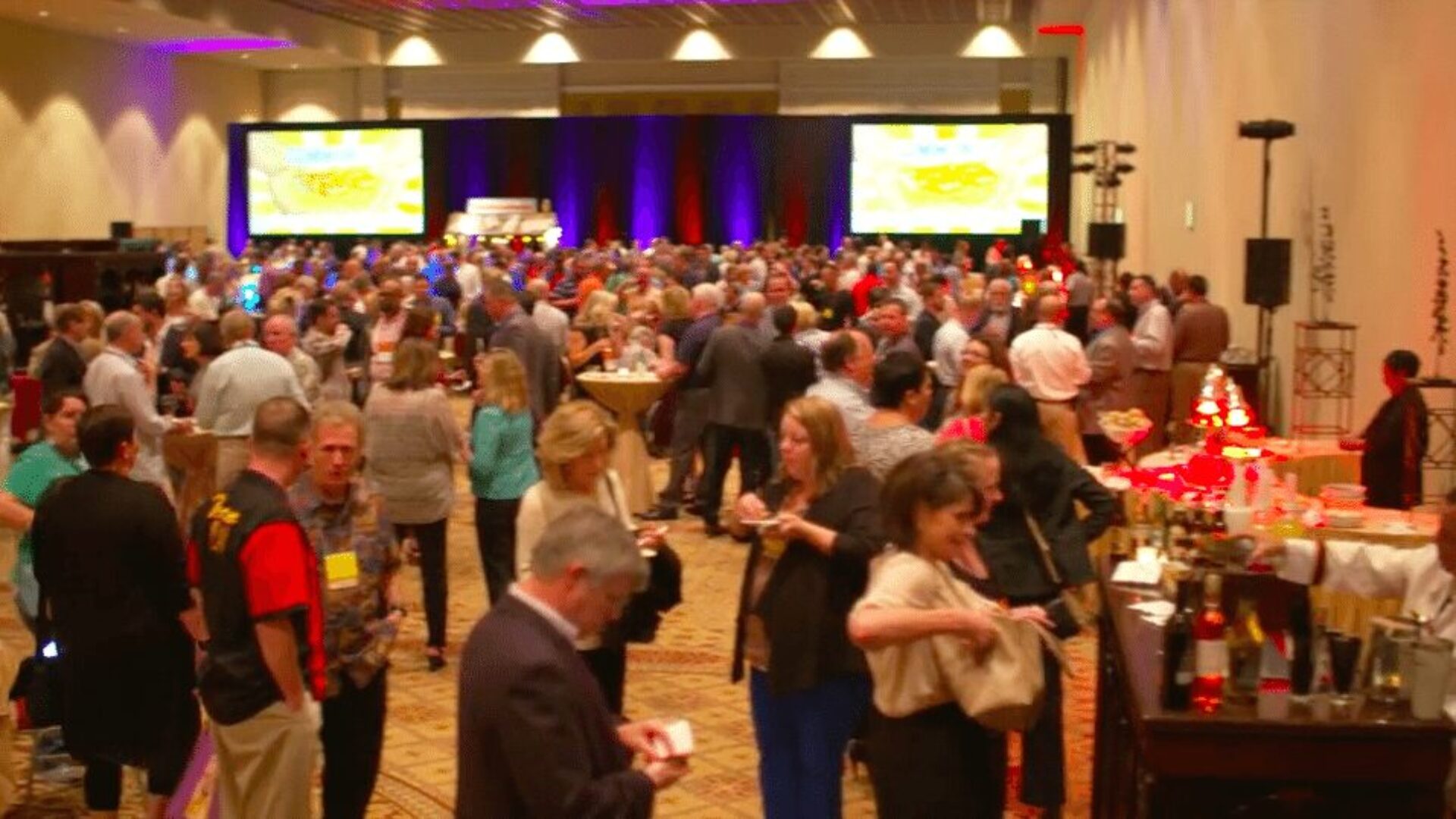 A crowded conference reception or networking event with attendees mingling in a large ballroom featuring stage screens and professional lighting.